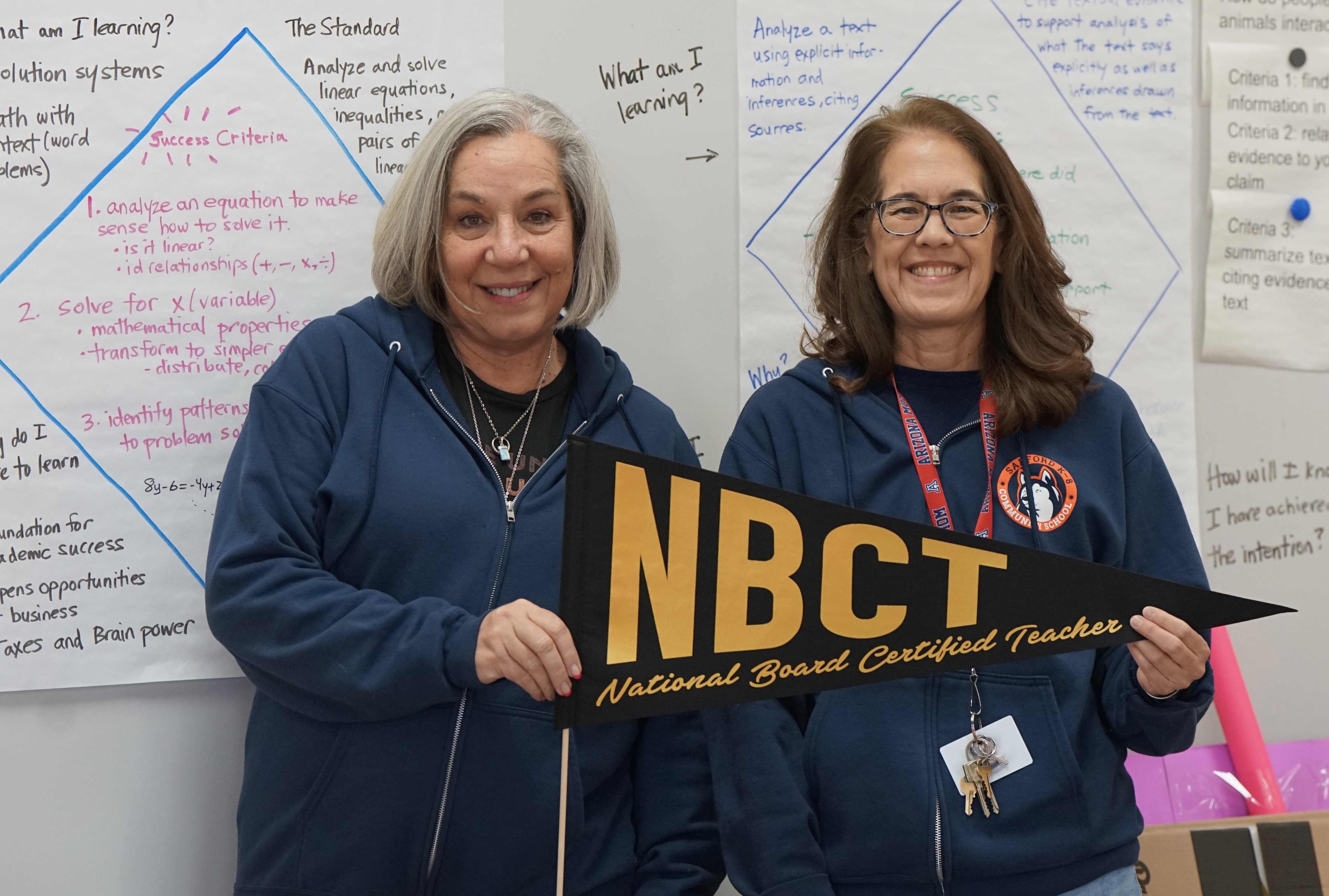 Two women in blue sweatshirts smile and hold an NBCT pennant